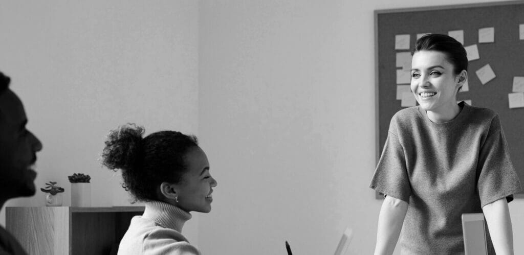 Smiling woman standing in front of bulletin board and talking to seated coworkers