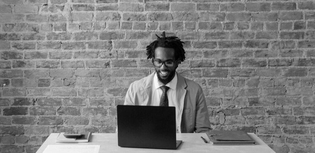 Man seated at a desk in front of a brick wall and typing at a laptop