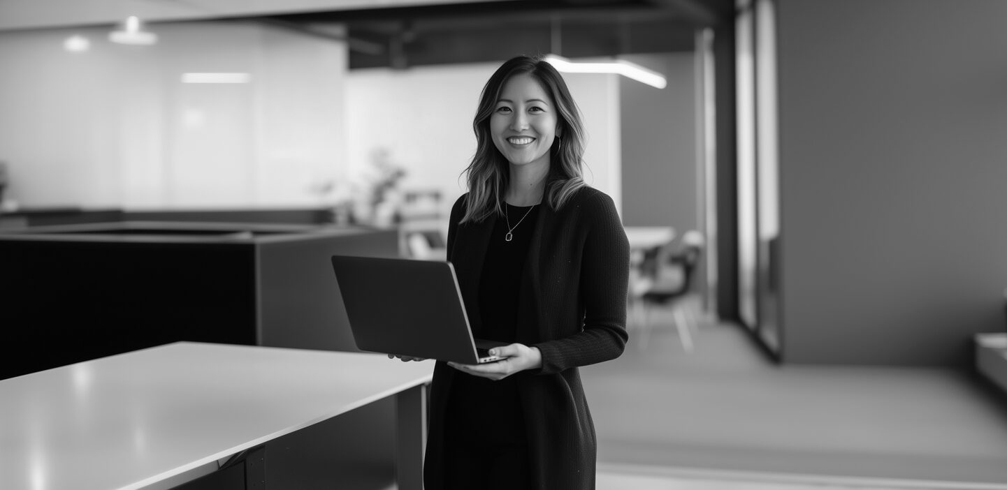 Woman standing in an office holds her laptop and smiles at the camera